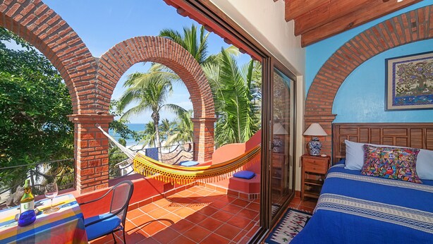 Una habitación con una cama, una hamaca, una mesa con una botella y un vaso, y vistas a palmeras y al mar.