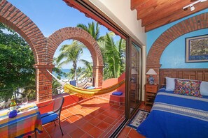 Une chambre avec un lit, un hamac, une table avec une bouteille et un verre, et une vue sur les palmiers et la mer.