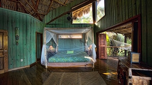 Una stanza con un letto coperte da zanzariera, pareti in legno, pavimento in legno, un'amaca e una finestra con vista sul verde.