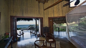 Una stanza in legno con un'ampia porta finestra che si apre su un balcone con vista sul mare. All'interno della stanza si trovano sedie e un tavolo in legno.