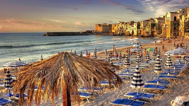 A beach with thatched umbrellas and sun loungers, a pier, and historic buildings along the shore.