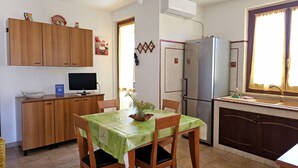 A compact kitchen with wooden cabinets, a refrigerator, a dining table with chairs, a small TV, and a window with curtains.