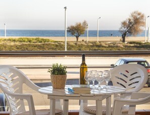 Une table avec une bouteille, des verres et un livre, donnant sur une plage où se trouvent quelques personnes et des arbres.