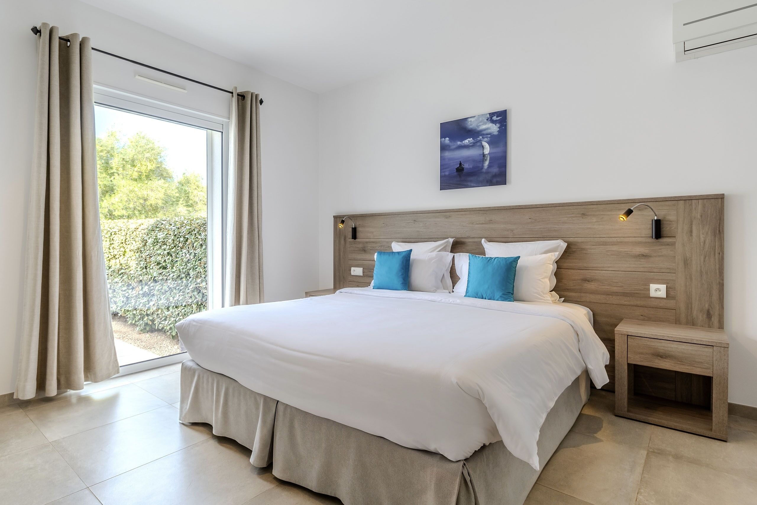 A bedroom with a large bed, a wooden headboard, a nightstand, and a view of greenery through a sliding door.