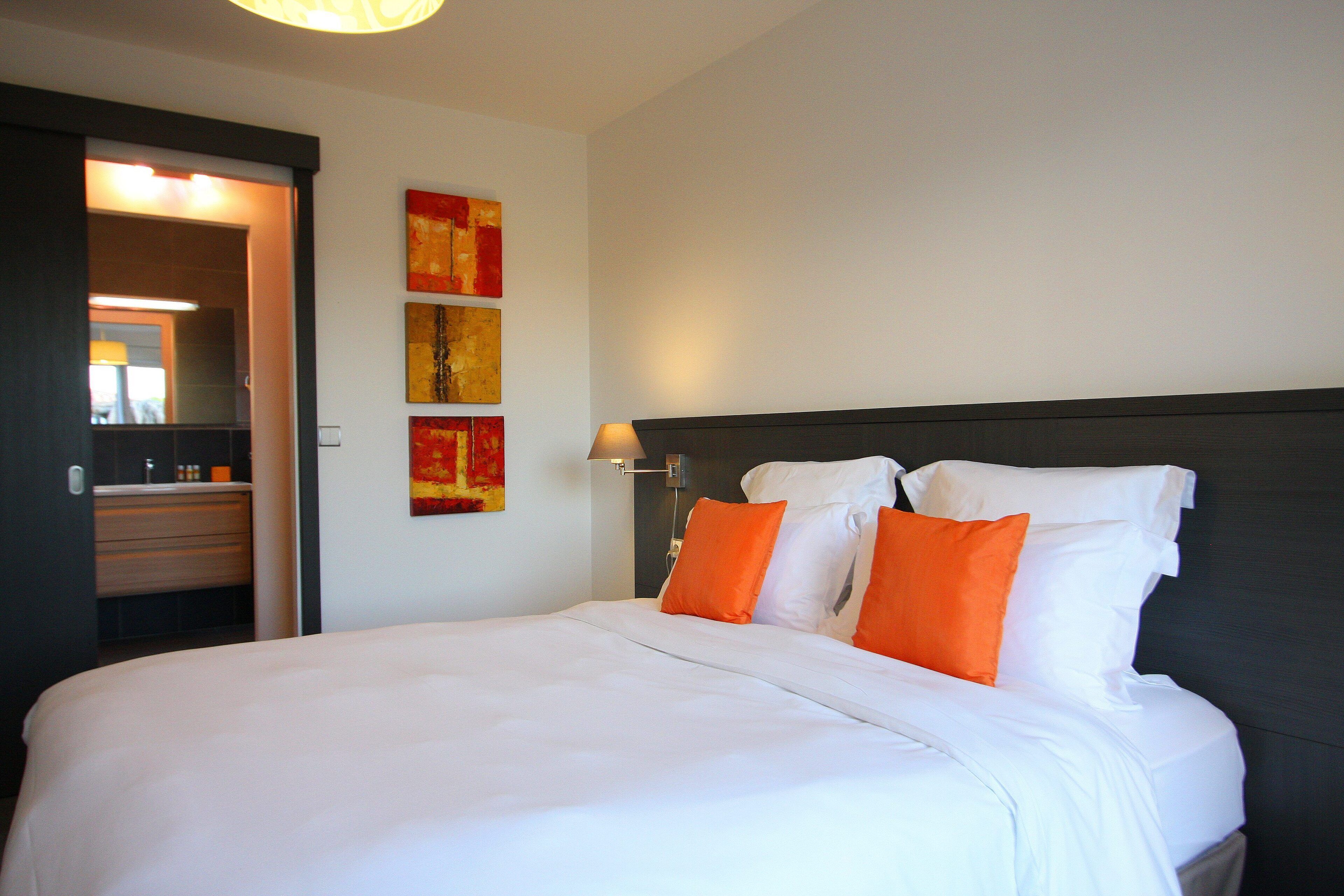 Ein Hotelzimmer mit einem großen Bett, weißer Bettwäsche und orangefarbenen Kissen. Ein Nachttisch mit Lampe und Spiegel sind vorhanden. An den Wänden befindet sich abstrakte Kunst.