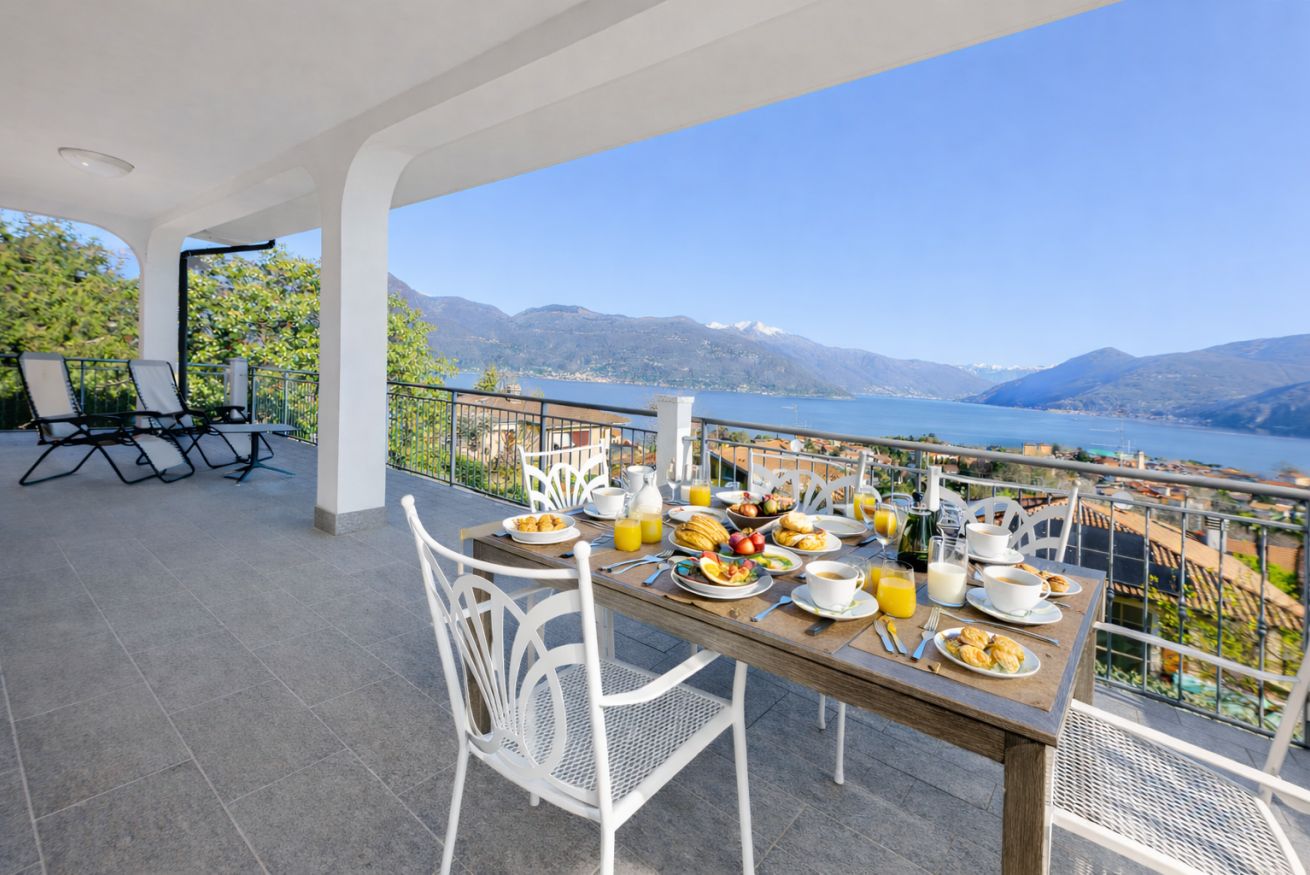 A terrace with a table set for breakfast, overlooking a lake and mountains.