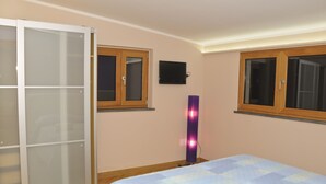Quarto com cama, guarda-roupa, duas janelas e televisão na parede.