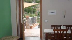 Quarto com um banco de madeira, uma mesa de jantar com cadeiras e vista para um pátio externo com parede de pedra e plantas em vasos.