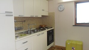 Uma cozinha compacta com armários brancos, pia, fogão e micro-ondas. Há um relógio na parede e uma janela que ilumina naturalmente o espaço.