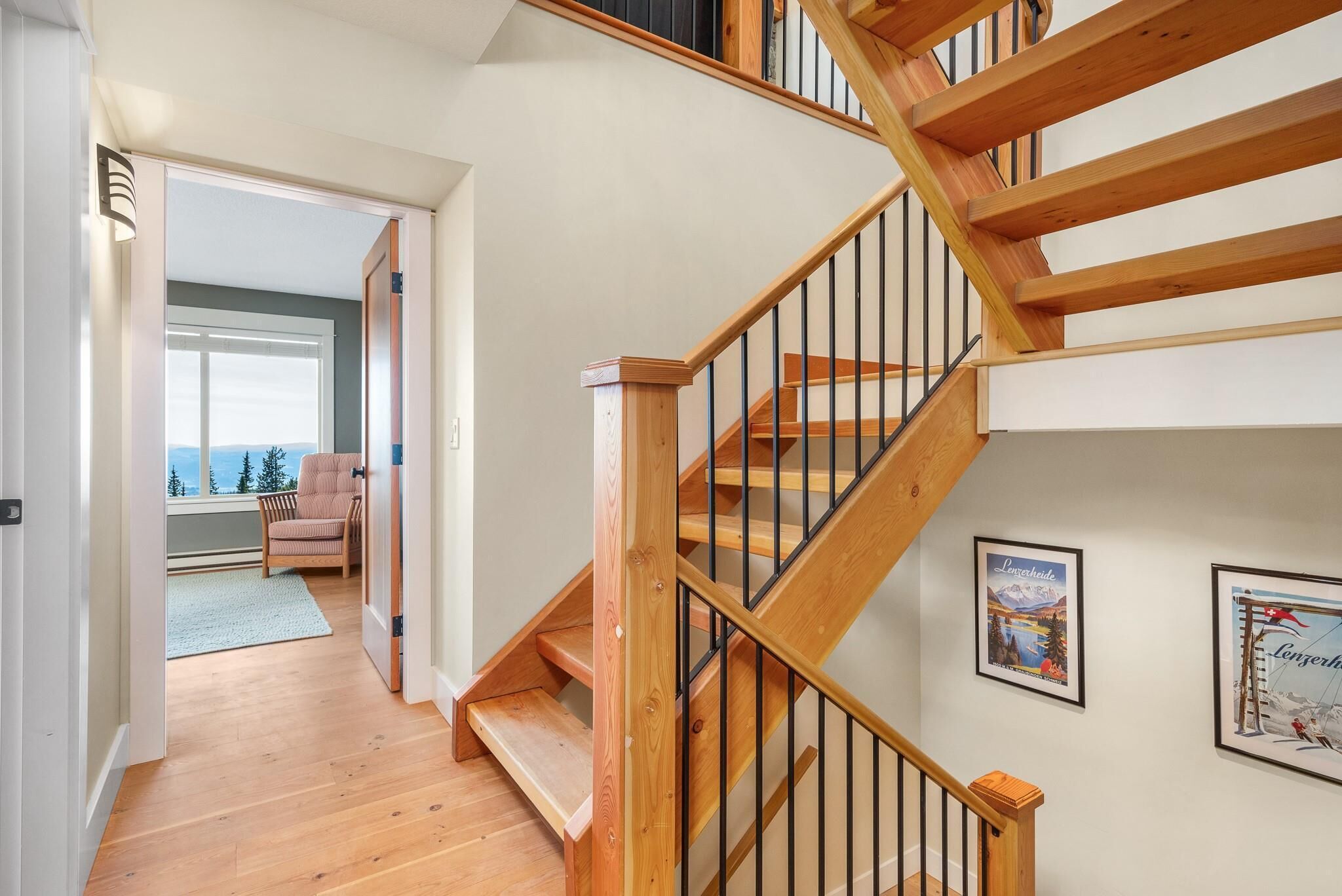 Eine Holztreppe mit schwarzen Geländern führt zu einer zweiten Etage. An den Wänden befinden sich gerahmte Bilder, im angrenzenden Raum steht ein Sessel und durch ein Fenster ist ein Blick ins Freie möglich.