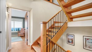 A wooden staircase with black railings leading to a second floor. There are framed pictures on the walls, a chair in the adjacent room, and a view of the outdoors through a window. - Upscale Ski In/Out Duplex on The Ridge with Garage, Private Hot Tub, laundry (Silver Star Mountain)