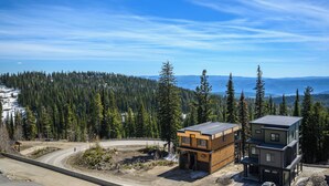 A modern two-story house with a flat roof, surrounded by a forested area and a view of mountains. - Upscale Ski In/Out Duplex on The Ridge with Garage, Private Hot Tub, laundry (Silver Star Mountain)