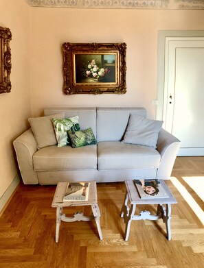 Uma sala de estar com um sofá cinza claro, duas mesinhas brancas, um quadro floral emoldurado e uma porta ao fundo.