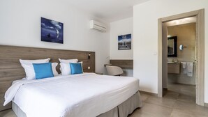 Une chambre d’hôtel avec un grand lit, une tête de lit en bois, un climatiseur fixé au mur et une salle de bain visible à travers une porte ouverte.