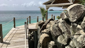Una terraza de madera que conduce a una construcción con techo de paja junto al mar, con grandes rocas en primer plano y una vista despejada del océano.