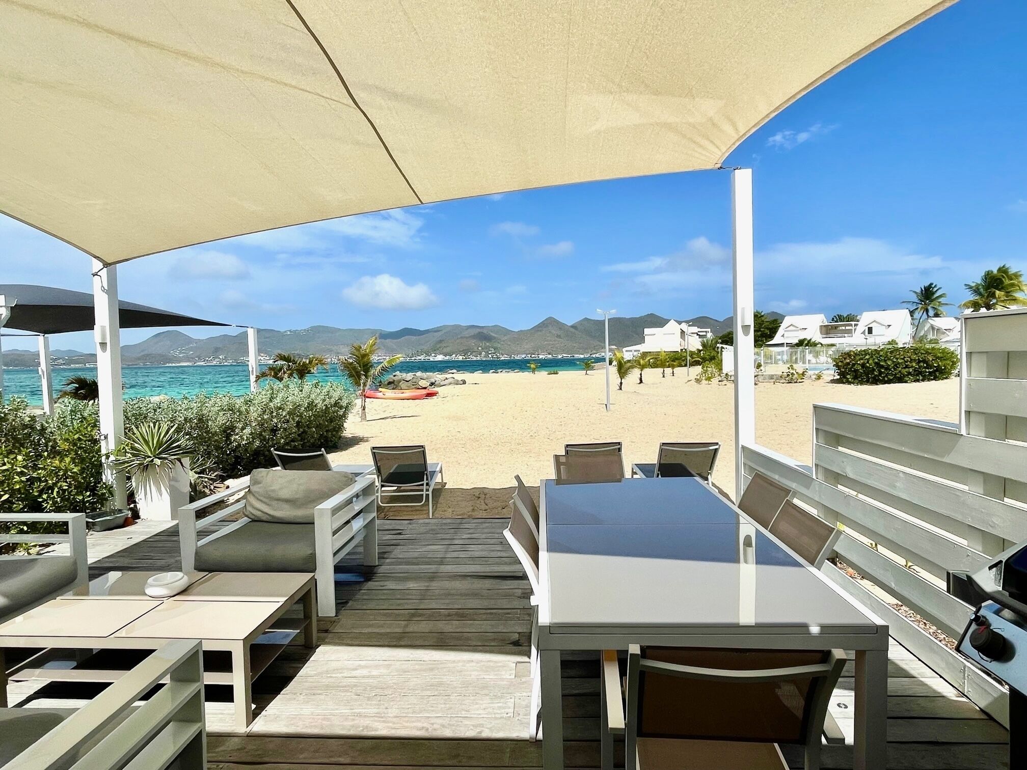 An outdoor seating area with tables and chairs, overlooking a beach with a clear view of the sea and distant mountains.