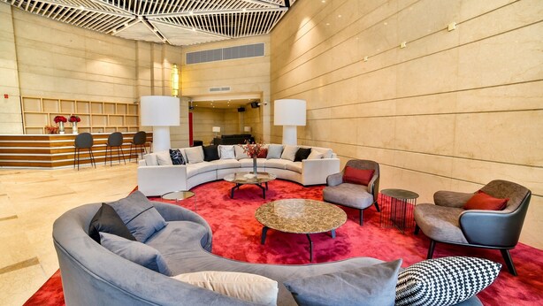 Un hall d’entrée spacieux, avec un grand canapé gris aux lignes courbes, une table basse ronde et un tapis rouge. On y trouve également deux fauteuils blancs et une petite table ronde surmontée d’un tapis rouge.