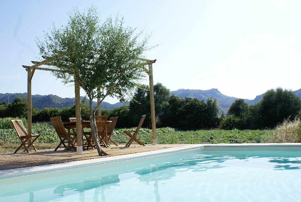 A pool area with wooden chairs and a table under a pergola, surrounded by greenery and mountains in the background.