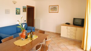 A dining area with a wooden table, chairs, a vase with flowers, a small table lamp, and a television on a cabinet. - Residence Garden: Three-room apartment with garden                            (Tignale)