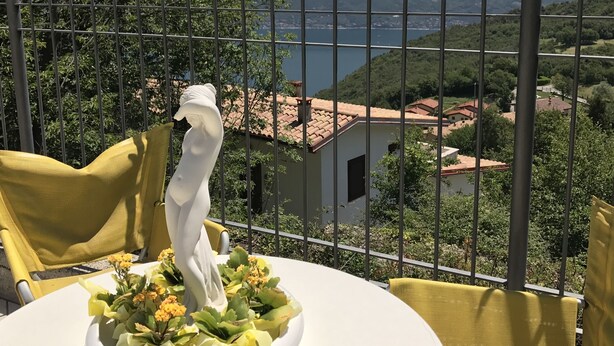 A balcony with a white table, yellow chairs, a white statue, and a view of mountains and a lake. - Residence Garden: Three-room apartment with garden                            (Tignale)
