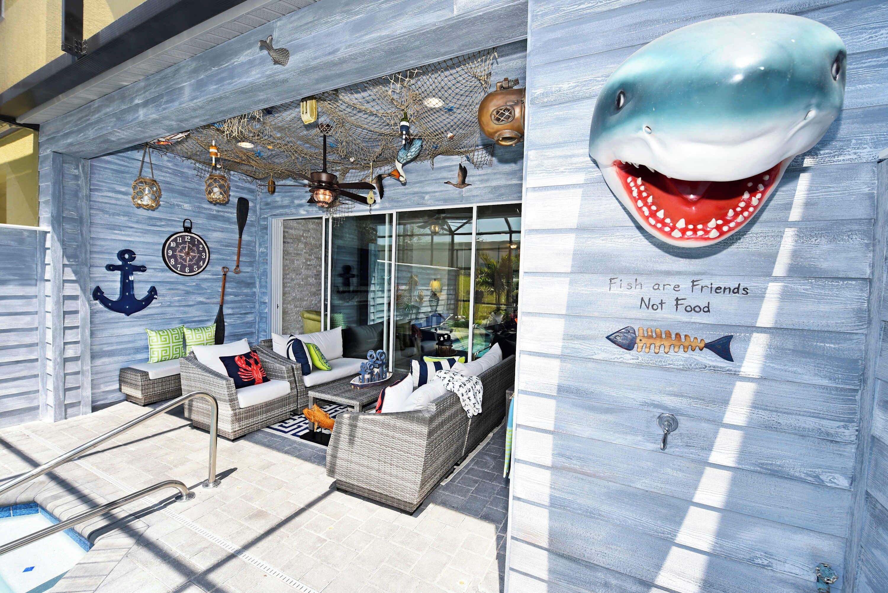 Un salón junto a la playa con asientos exteriores, piscina y una pared decorativa con una ilustración de tiburón y el texto "Fish are Friends Not Food".