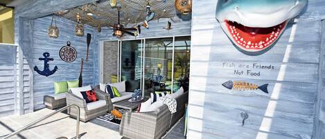 Un salón junto a la playa con asientos exteriores, piscina y una pared decorativa con una ilustración de tiburón y el texto "Fish are Friends Not Food".