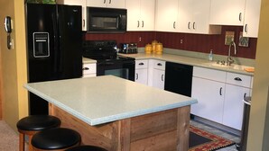 Une cuisine compacte avec des meubles blancs, un réfrigérateur noir, un four à micro-ondes, une cuisinière et un évier. Il y a trois tabourets en bois près de l’îlot de cuisine.