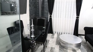 Een moderne kamer met een glazen eettafel, zwarte beklede stoelen, een zwarte bank, een rond poef, een zwart haardvuur en een grote spiegel.