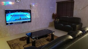 A living room with a flat-screen TV displaying a tennis game, a black leather sofa, and a coffee table. - Grand Valley Hotel (Abuja)