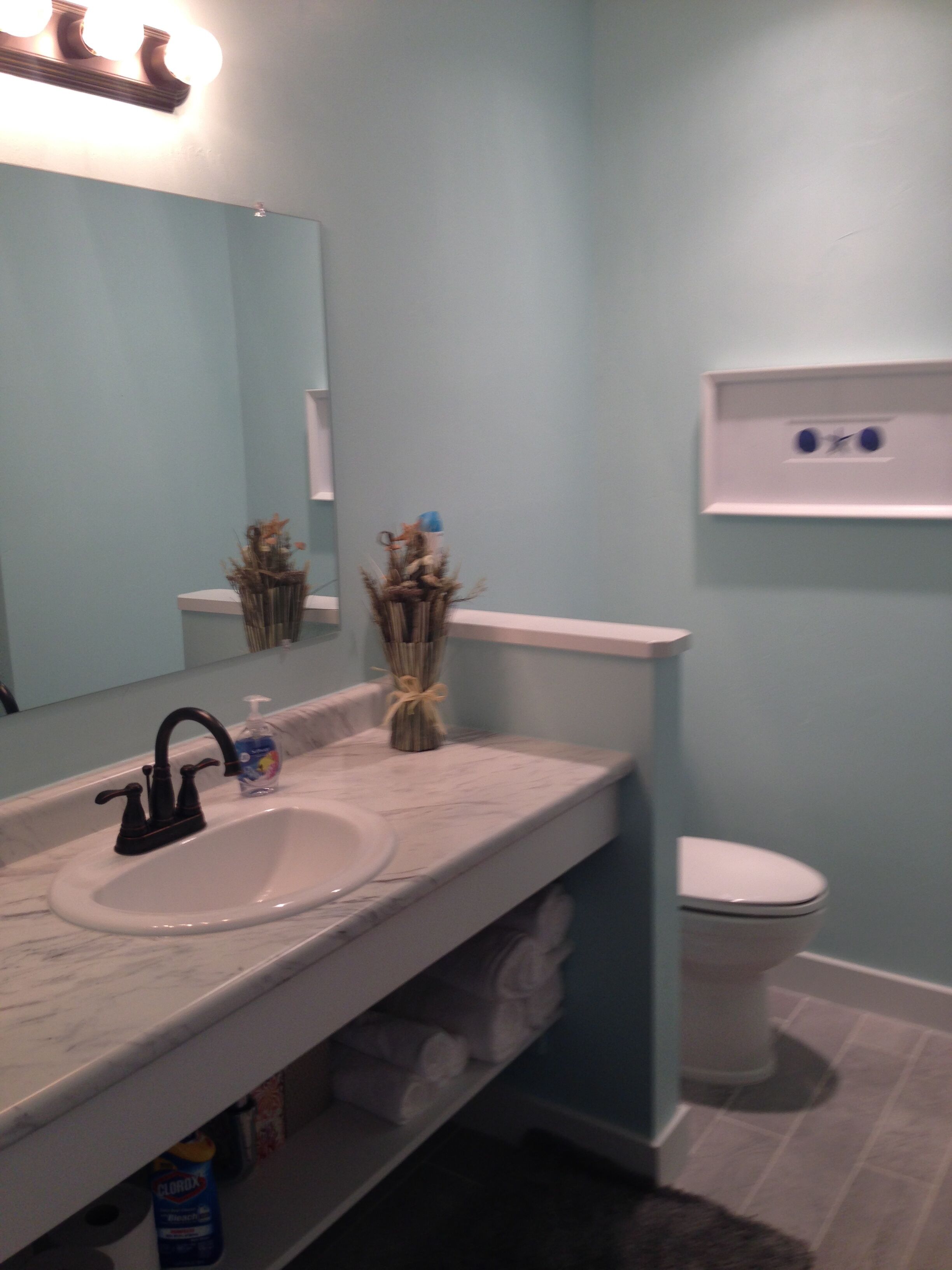 Een badkamer met een witte wasbak, een zwarte kraan en een spiegel erboven. Rechts bevindt zich een toilet en onder de wasbak een plank met handdoeken.