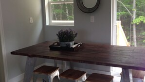 Un coin repas avec une table en bois, trois tabourets en métal, une plante en pot et une fenêtre donnant sur des arbres.