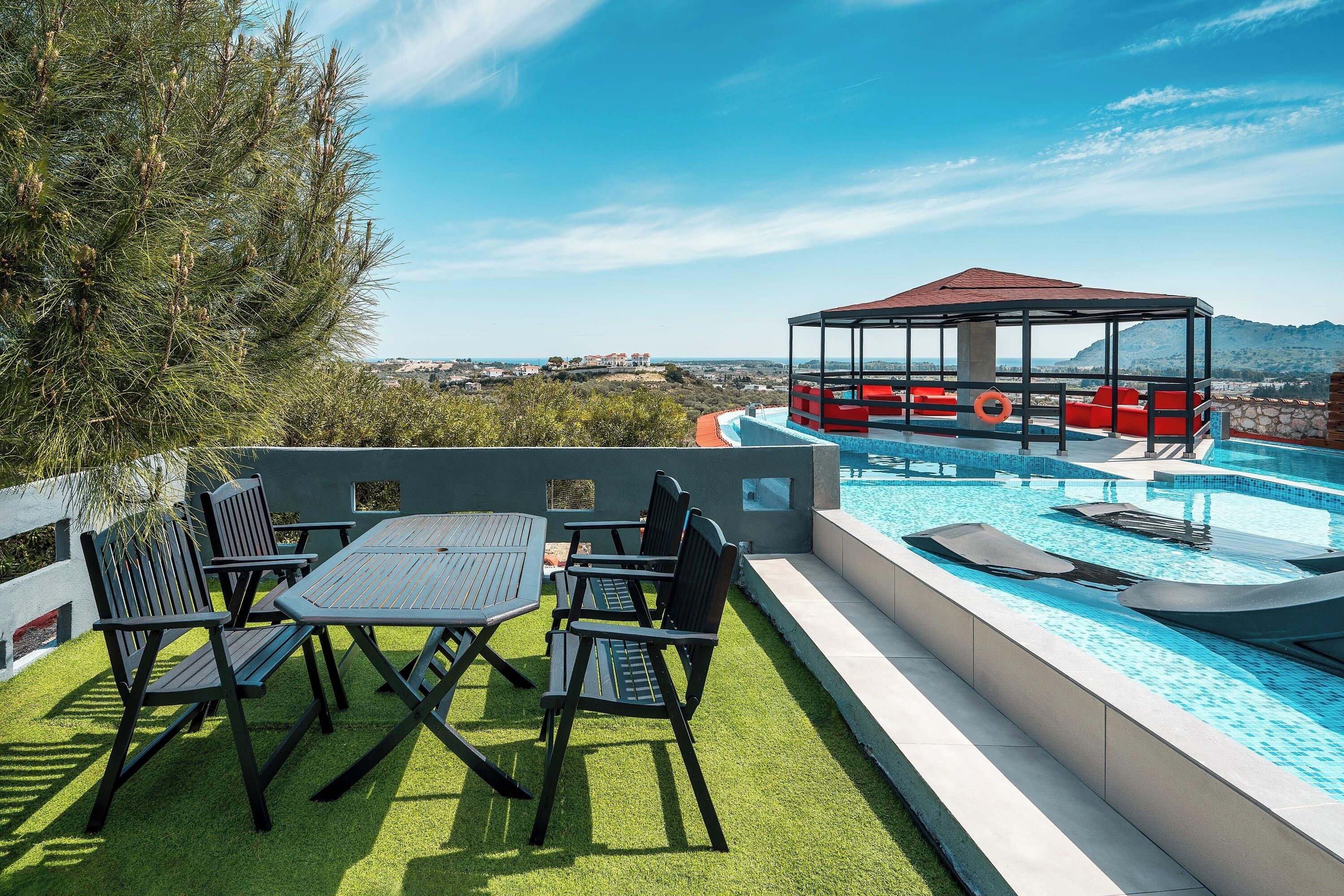 An outdoor pool area with a covered seating area, a table with chairs, and a view of the landscape.