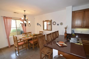 Un coin repas avec des chaises et une table en bois, une cuisine avec des meubles en bois, un lustre et une fenêtre avec des rideaux.