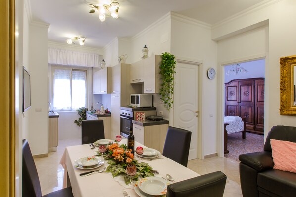 Un coin repas avec une table dressée pour quatre personnes, une cuisine en arrière-plan et un salon avec un canapé et un miroir.
