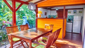 A wooden dining table with chairs, a kitchen area with a refrigerator and a coffee maker, and a view of greenery outside.
