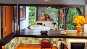 A kitchen with a wooden countertop, a red toaster, a kettle, and a microwave. There is a hanging light fixture above the counter and a window with a view of a garden.