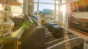 A modern living room with a sofa, ottomans, a fireplace, and large windows offering a city view. - EraBella-Downtown 12th floor Beauty, Free Parking and Wifi, Close to Everything by Victoria Prime! (Victoria)
