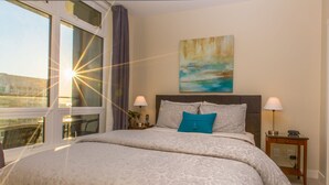 A spacious bedroom with a large bed, two bedside lamps, a nightstand, a painting on the wall, and a view of the outside through a window. - EraBella-Downtown 12th floor Beauty, Free Parking and Wifi, Close to Everything by Victoria Prime! (Victoria)