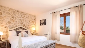 A bedroom with a stone wall, a bed with a bench, two bedside tables with lamps, a window with curtains, and a picture on the wall.
