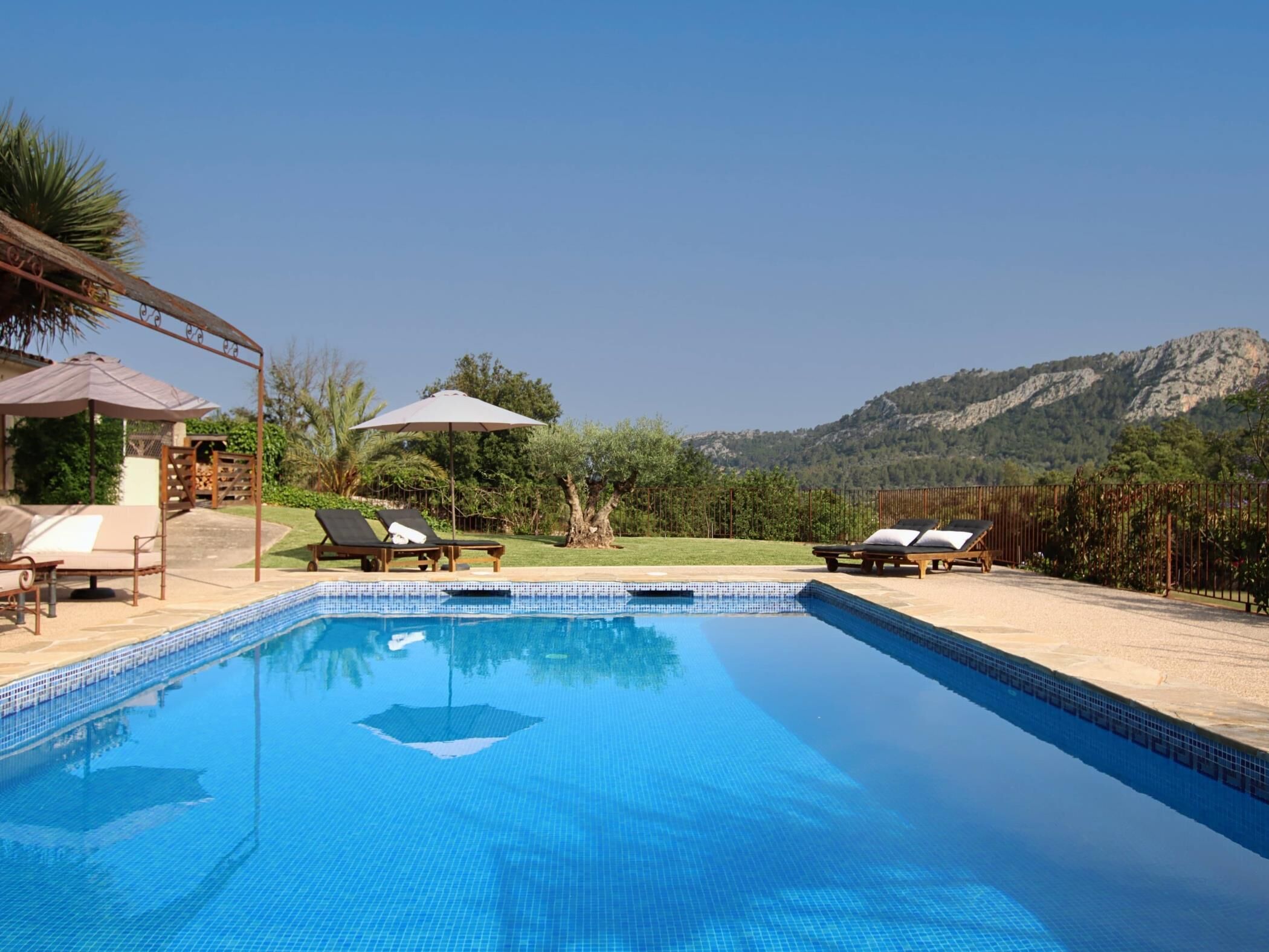 Ein Schwimmbecken mit Liegestühlen und Sonnenschirmen, umgeben von einem angelegten Garten und Bergen im Hintergrund.