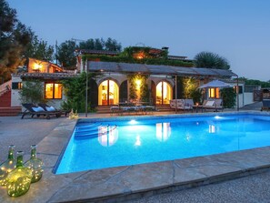 A poolside area with a swimming pool, lounge chairs, and a building with arched windows and outdoor lighting.