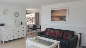 A living room with a sofa, a coffee table, a dining area with a table and chairs, a kitchen area with a stove, a clock, and a painting on the wall. - Amazing panoramic sea views!!!  (Playa Blanca)