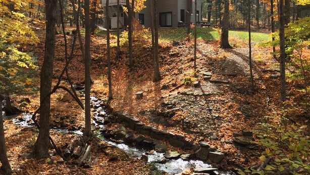 A cabin surrounded by autumn foliage, a small stream, and a grassy area.