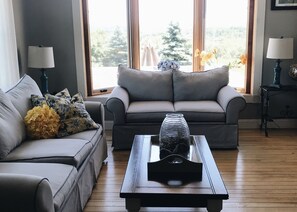 Ein Wohnzimmer mit grauem Sofa, einem Holztisch und einem Fenster mit Blick auf Bäume.