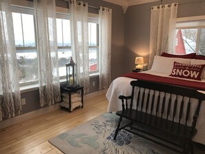 Ein Schlafzimmer mit einem Bett, einem Nachttisch, einer Lampe, einem Sessel und Blick durch große Fenster nach draußen.
