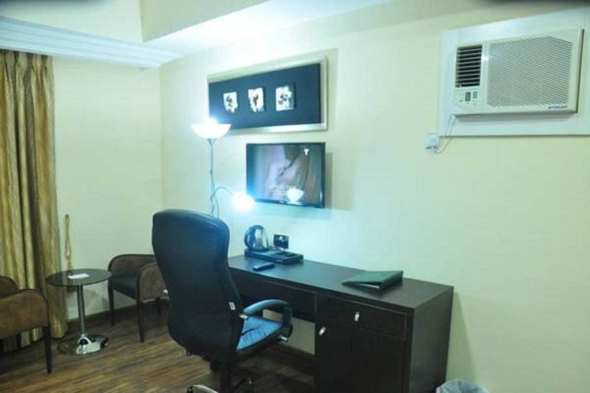 Een hotelkamer met een bureau, een stoel, een lamp, een klein tafeltje, een televisie en een airconditioner.