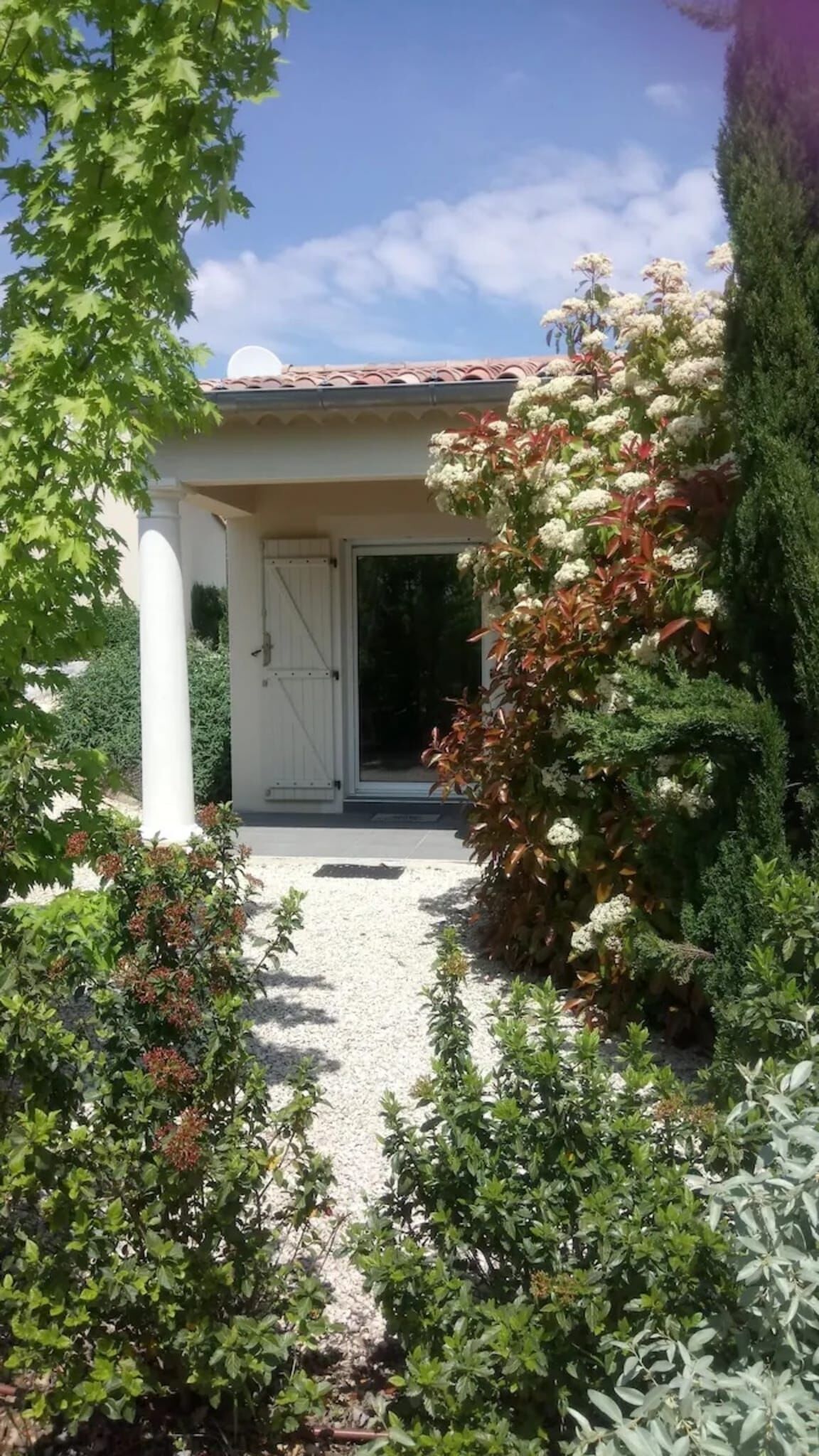 Un espace extérieur abrité, doté d’un chemin de gravier menant à une porte, et entouré de divers arbustes et plantes.