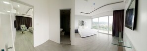 Una habitación de hotel moderna con una cama grande, una mesa de vidrio, un televisor montado en la pared y vistas de la ciudad a través de amplias ventanas.
