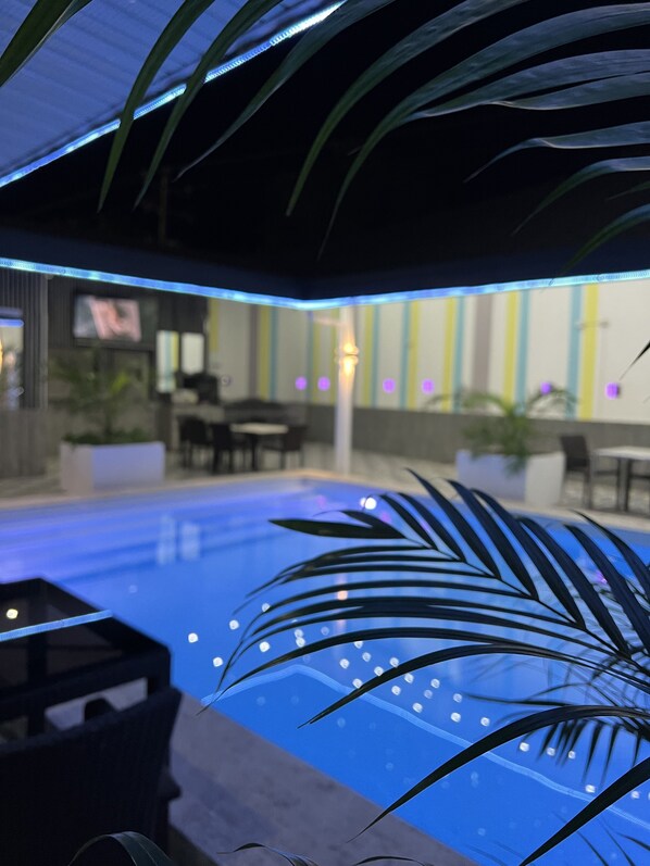 Un área de piscina interior con una piscina azul, asientos alrededor de la piscina e iluminación azul en el techo.