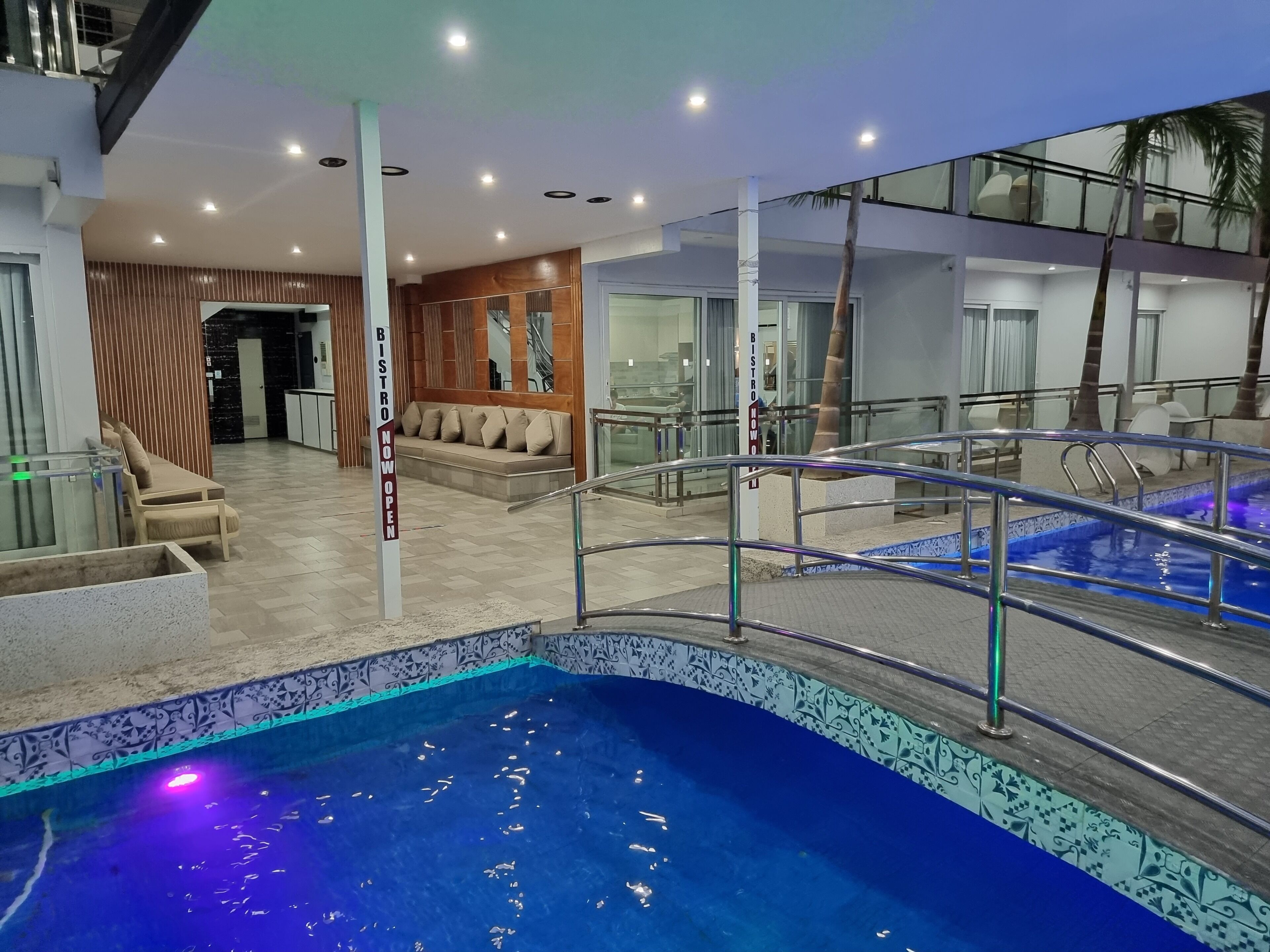 A modern indoor pool area with a glass railing, a seating area with a 'BISTRO NOW OPEN' sign, and a view of the adjacent restaurant.